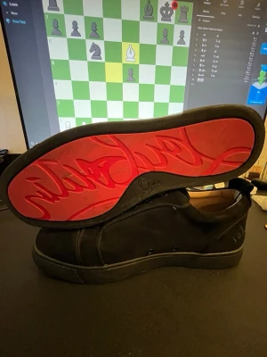 Svarta Louboutin sneakers med röd sula - Stilrena svarta sneakers från Christian Louboutin i mocka med klassisk röd sula och diskret logga på sidan. Lågt skaft och svarta skosnören ger en clean look. Perfekta för dig som vill sticka ut med exklusiva detaljer.