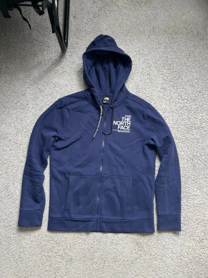 Blå hoodie från The North Face - Snygg blå hoodie från The North Face med dragkedja framtill och två stora fickor. Klassisk logga i vitt på bröstet och ryggen. Tillverkad i mjukt bomullsmaterial och har justerbar huva med snören. Perfekt för en avslappnad streetwear-look.
