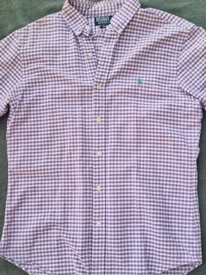 Lila rutig skjorta från Ralph Lauren - Snygg lila och vit rutig skjorta från Ralph Lauren med klassisk krage och knäppning framtill. Skjortan har långa ärmar och den ikoniska gröna broderade loggan på bröstet. Perfekt för en stilren och avslappnad look.