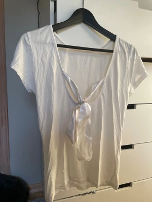 Vit topp med knyt i ryggen från Lager 157 - Vit topp med urringning i ryggen med knyte. Den sitter ganska tajt i midjan och har ett tunnare material. Den är i L men är liten i storlek och sitter tajt även om man brukar ha M/S. 