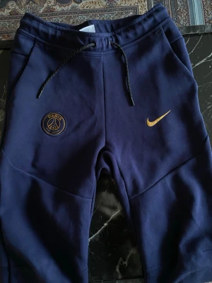 Blå PSG x Nike mjukisbyxor - Snygga mörkblå mjukisbyxor från Nike i samarbete med PSG. Byxorna har guldfärgade detaljer med PSG-logga och Nike Swoosh på framsidan. Utrustade med dragsko i midjan, fickor på sidorna och muddar vid bensluten. Perfekta för chill eller sportiga dagar.