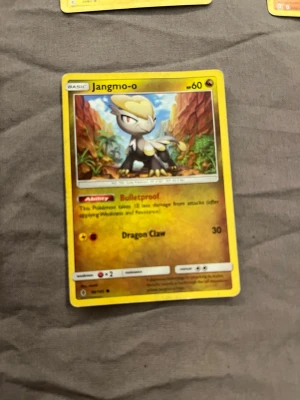 Pokémonkort Jangmo-o - Säljer ett Pokémonkort med Jangmo-o, gul ram och färgglad illustration. Kortet har förmågan Bulletproof och attacken Dragon Claw. Perfekt för samlare eller Pokémonfans som vill utöka sin kortlek.