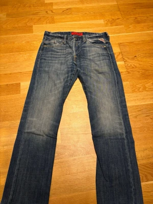 Blå raka jeans från Replay - Säljer ett par klassiska blå jeans från Replay i modellen Waitom. Jeansen har raka ben, fem fickor och snygga slitningar för en avslappnad look från fabrik. Tillverkade i slitstark denim med detaljerade sömmar och röd Replay-logga på fickan. Strl 31/32