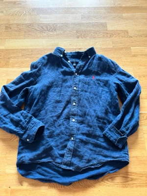 Navy linneskjorta från Polo Ralph Lauren - Säljer en blå långärmad skjorta i linne från Polo Ralph Lauren med klassisk button-down krage och röd broderad logga på bröstet. Skjortan har vita knappar och en avslappnad passform, perfekt för en cool och stilren look. Storleken är 14-16 år gammal 👍den är i fint skick✅