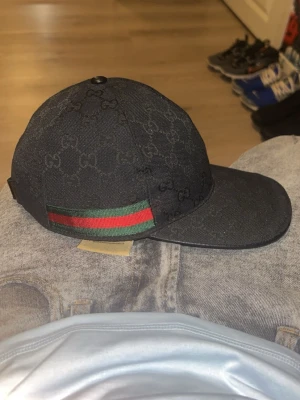 Svart Gucci keps med GG-mönster - Svart keps från Gucci med klassiskt GG-mönster över hela kepsen. På sidan finns en grön och röd rand som ger kepsen en lyxig touch. Materialet är canvas och kepsen har böjd skärm. Perfekt för dig som vill ha en exklusiv accessoar storlek M 58cm bra skick inga fel med den 10,10✅✅