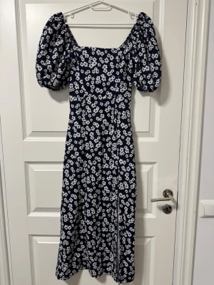 Marinblå blommig långklänning - Snygg marinblå långklänning med vita blommor och puffiga kortärmar. Klänningen har en fyrkantig ringning och en smickrande passform. 