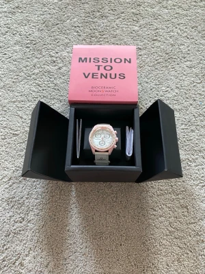 Omega x Swatch Mission to Venus klocka - Säljer en Omega x Swatch Mission to Venus från Bioceramic MoonSwatch Collection. Klockan har en ljusrosa boett, vit urtavla med kronografer och vit rem med Omega-logga. Snygg och unik design som sticker ut på handleden.