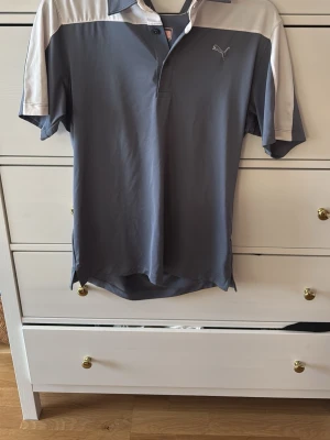 Grå sportpiké från Puma - Snygg grå sportpiké från Puma med vita axelpartier och klassisk krage. Tröjan har korta ärmar och är tillverkad i ett lätt och funktionellt material som andas, perfekt för träning eller sport. Puma-logga broderad på bröstet.