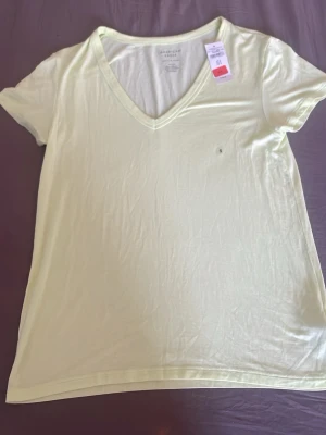 Ljusgul v-ringad t-shirt American Eagle - Fräsch ljusgul t-shirt från American Eagle med v-ringning och korta ärmar. Tillverkad i mjuk bomullsmix som känns skön mot huden. Enkel och stilren design utan mönster, perfekt till jeans eller shorts.