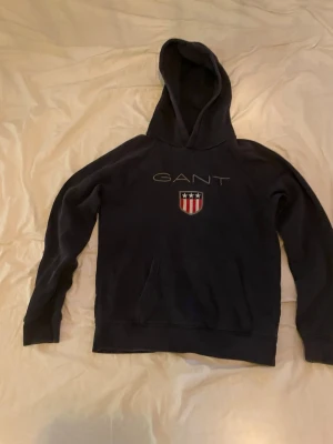 Mörkblå hoodie från GANT - Snygg mörkblå hoodie från GANT med broderad logga och sköld med stjärnor och ränder på bröstet. Klassisk känguruficka framtill och stor huva. Perfekt för en avslappnad och sportig stil.