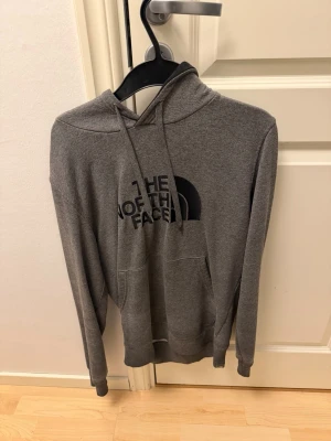 Grå hoodie från The North Face - Snygg grå hoodie från The North Face med stor logga på bröstet. Klassisk modell med huva och dragsnören samt en stor magficka. Tillverkad i mjukt material som känns skönt mot huden. Perfekt för en avslappnad och cool stil.