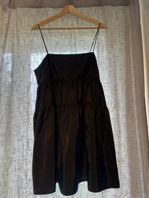 Svart kort klänning med tunna axelband - Svart kort klänning med tunna axelband och voluminös, luftig passform. Klänningen har flera volangliknande sömmar.