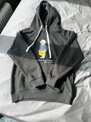 Svart Polo Bear hoodie från Ralph Lauren - Säljer en svart hoodie från Ralph Lauren med den ikoniska Polo Bear-trycket framtill. Hoodien har vita dragsnören, känguruficka och ribbade muddar. Materialet känns mjukt och bekvämt, perfekt för dagar där du vill klä dig grisch utan att tappa bekvämligheten.
