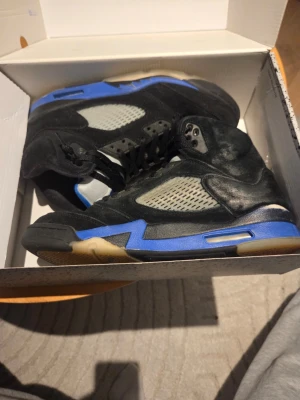 Air Jordan 5 Retro svart/blå - Säljer ett par Air Jordan 5 Retro sneakers i svart mocka med blå och grå detaljer. Skorna har meshpaneler på sidorna, klassisk hög siluett och synlig Air-enhet i sulan. Perfekta för dig som gillar streetwear och ikoniska sneakers.