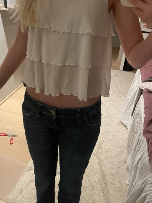 Vit volangig linnetopp med smala band - Säljer en söt vit linnetopp med smala axelband och flera lager volanger. Toppen är croppad och har en luftig känsla, perfekt för varma dagar. Enkel att matcha med jeans eller kjol för en trendig look.