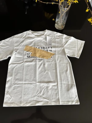 Maison Margiela t-shirt - Säljer en vit t-shirt från Maison Margiela med ikoniskt siffertryck och beige tejpdetalj över bröstet. T-shirten har rund hals, korta ärmar och är gjord i mjuk bomull. Perfekt för dig som gillar unika designer och streetwear vibes.