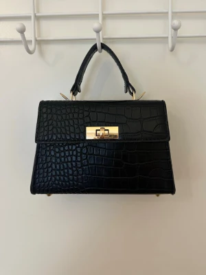 Svart handväska med krokodilmönster - Stilren svart handväska i konstläder med krokodilmönstrad yta och guldfärgade metalldetaljer. Väskan har ett topphandtag och ett elegant vridlås framtill. Perfekt accessoar för att lyfta din outfit med en trendig touch🫶🏼