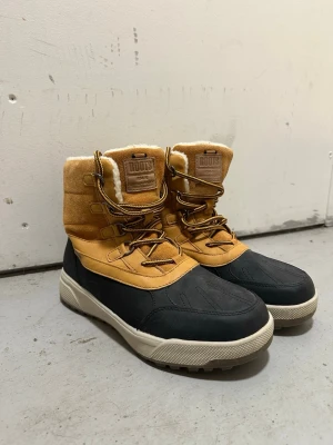 Fodrade vinterboots från Roots - Säljer ett par snygga vinterboots från Roots med beige mocka upptill, svart vattentålig nederdel och grov sula. Bootsens insida är fodrad med mjuk pälsimitation och de har klassiska snörningar med tvåfärgade skosnören. Perfekta för kalla dagar och outdoor vibes.