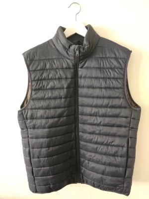 Svart dunväst med ståkrage - Snygg svart dunväst med ståkrage och dragkedja framtill. Västen har ett quiltat mönster och är lätt vadderad för extra värme. Perfekt att slänga över en hoodie eller tröja för en chill look.
