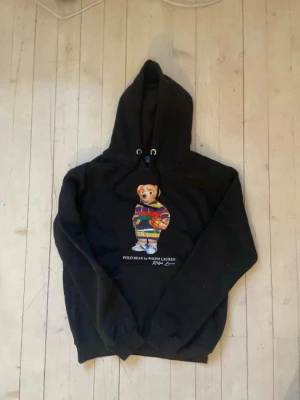 Svart hoodie Polo Bear Ralph Lauren - Svart hoodie från Ralph Lauren med den ikoniska Polo Bear-trycket på bröstet. Björnen har färgglada kläder och håller en basketboll. Hoodien har dragsko i huvan och ribbade muddar. Perfekt för dig som vill ha en streetwear-vibe med en lekfull twist. Säljer pga fel storlek, pris kan diskuteras i snabb affär😁