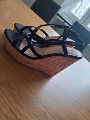 Svarta kilklackssandaler från H&M - Snygga svarta sandaler från H&M med remmar och hög kilklack i korkimitation. Perfekta för dig som vill ha både höjd och komfort. Ovandelen är i svart tyg och sulan är ljus med en naturlig look. Stängs med rem runt vristen.