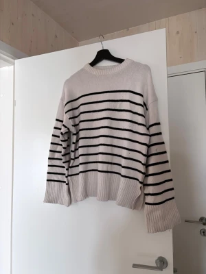 Randig stickad tröja i offwhite - Säljer en stickad tröja i offwhite med svarta horisontella ränder. Tröjan har rund halsringning, långa ärmar och breda ribbstickade muddar vid ärmslut och nederkant. Oversized passform för en avslappnad look.