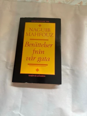 Berättelser från vår gata - Mahfouz - En skönlitterär bok av Naguib Mahfouz med titeln 'Berättelser från vår gata'. Omslaget är svart med en gul och röd rektangel där titeln och författarens namn står. Boken är i pocketformat och tryckt på papper.
