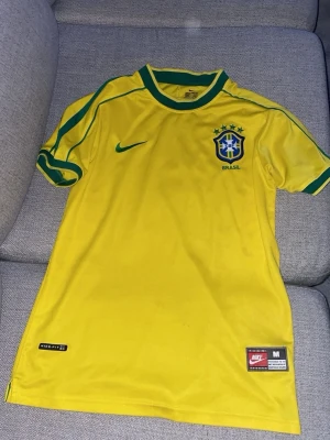 Gul Brasilien Nike fotbollströja - Säljer en klassisk gul Brasilien fotbollströja från Nike med gröna detaljer på ärmar och krage. Tröjan har Brasilien-emblemet på bröstet och Nike-logga i grönt. Tillverkad i lätt och ventilerande material, perfekt för träning eller match.