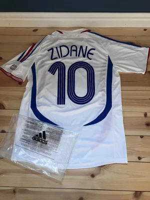 Frankrike Zidane tröja Adidas - Säljer en vit fotbollströja från Adidas med Zidane och nummer 10 på ryggen. Tröjan har blå och röda detaljer, Frankrikes landslagsmärke på bröstet och Adidas-logga. Korta ärmar med ränder i blått och rött. Perfekt för dig som älskar fotboll och klassiska landslagströjor.