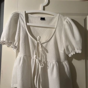 Vit kort klänning från Gina Tricot - Supersöt vit kort klänning från Gina Tricot med puffärmar och knyt framtill. Klänningen har en lätt rynkad struktur och en feminin siluett med volangdetaljer vid ärmslut. Perfekt för varma dagar och somriga tillfällen.