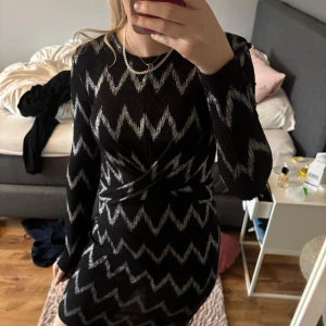 Svart och silverglittrig klänning - Snygg svart kortklänning med silvrigt glittrigt zigzag-mönster. Klänningen har lång ärm, omlott-detalj framtill och en öppen rygg. Perfekt för dig som vill sticka ut med en cool look.