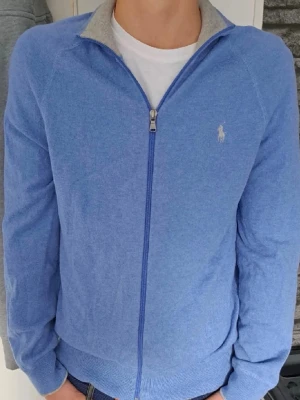 Blå ziptröja från Polo Ralph Lauren - Snygg blå ziptröja från Polo Ralph Lauren i storlek M. Tröjan har hel dragkedja framtill, ribbade muddar och är tillverkad i mjukt material som känns skönt mot huden. Perfekt för att slänga på sig när det är lite kyligt ute.
