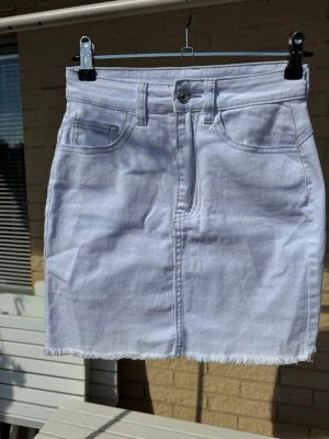 XS Vit jeanskjol med rå kant - Snygg vit jeanskjol med klassisk femficksdesign och rå, fransad nederkant. Kjolen har knapp och dragkedja framtill samt bälteshällor. 🤍