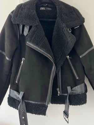 Svart teddyjacka från Zara med detaljer  - Snygg svart teddyjacka från Zara med stora slag, dragkedjor och bältesdetaljer. Jackan har mjukt teddyfoder, detaljer i fuskskinn och flera fickor med dragkedja. Perfekt för kalla dagar och ger en cool look.