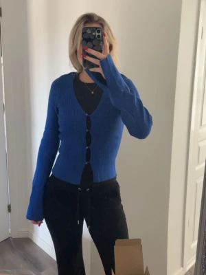 Kornblå ribbad kofta med knappar - Säljer en kornblå ribbad kofta med lång ärm och djup v-ringning. Koftan har fyra matchande knappar framtill och är figurnära i modellen. Perfekt att styla över ett linne eller t-shirt för en trendig look.