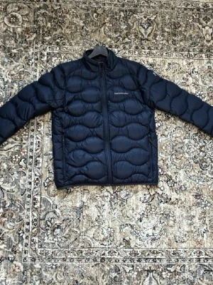 Mörkblå pufferjacka från Peak Performance - Snygg mörkblå pufferjacka från Peak Performance med vågformad quiltning och hög krage. Jackan har dragkedja framtill och är tillverkad i ett glansigt material som ger en modern look. Perfekt för kalla vinterdagar. Pris kan ej diskuteras 