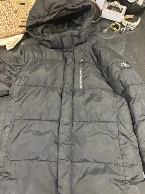 Svart pufferjacka från Calvin Klein - Snygg svart pufferjacka från Calvin Klein Jeans med huva och dragkedja framtill. Jackan har en diskret logga på bröstet och ärmen samt stor logotyp på insidan. Tillverkad i glansigt, vadderat material som håller dig varm under kalla dagar.