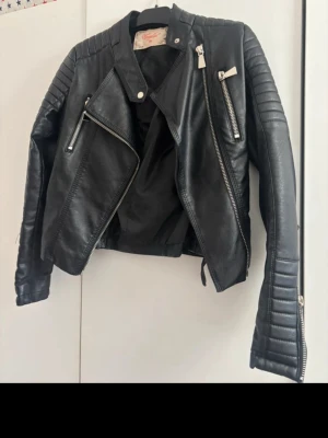 Svart biker skinnjacka med dragkedjor - Snygg svart biker skinnjacka med coola detaljer som silvriga dragkedjor, quiltade partier på axlar och ärmar samt snedställd dragkedja framtill. Jackan har en klassisk krage och figurnära passform. 
