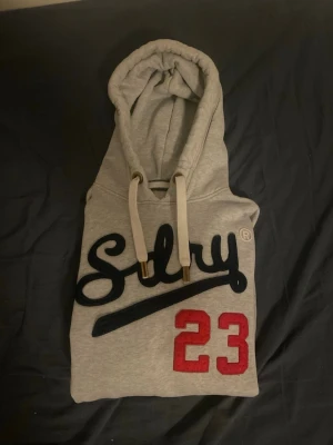 Grå hoodie från Superdry med tryck - Snygg grå hoodie från Superdry med stor huva och vita dragsnören. Framtill står det 'Sdry' i mörkblått och siffran 23 i rött. Känguruficka och patch vid nederkanten. Perfekt för en avslappnad streetwear-look.