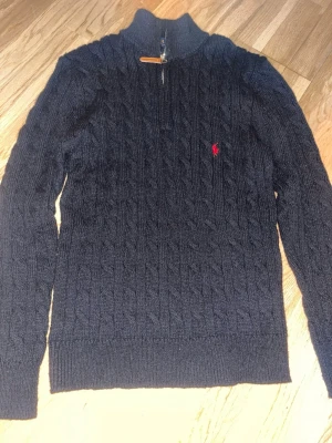 Mörkblå kabelstickad tröja Polo Ralph Lauren - Kabelstickad mörkblå tröja från Polo Ralph Lauren med halv dragkedja och röd broderad logga på bröstet. Tröjan har hög krage och ribbade muddar. Perfekt för lager-på-lager och snygg till jeans.Half zip stilig och passar som vardaglig still och lite finare. Pris kan diskuteras 
