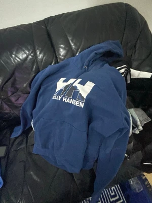 Blå Helly Hansen hoodie med tryck - Snygg blå hoodie från Helly Hansen med stort vitt och mörkblått logotryck på bröstet. Klassisk modell med huva och känguruficka framtill. Perfekt för en avslappnad och sportig stil.