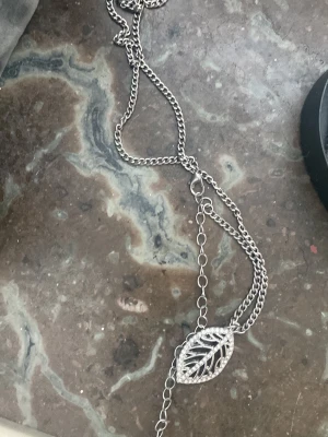 Silverfärgat halsband med bladberlock - Snyggt halsband i silverfärgad metall med flera kedjor och ett stort bladformat hänge. Hänget har ett detaljerat, öppet mönster som ger en modern och trendig look. Perfekt accessoar för att lyfta din outfit med en unik touch.