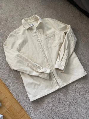 Weekday Overshirt - Säljer en beige skjortjacka från Weekday i storlek S. Skjortan har lång ärm, klassisk krage, bröstficka och knappar framtill. Materialet är mjukt och ger en avslappnad men ändå stilren look. Perfekt att bära öppen eller stängd.