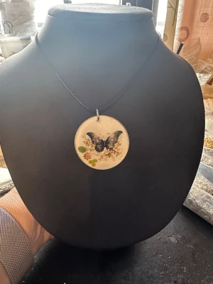 Halsband med fjäril och blommor - Unikt halsband med runt hänge i transparent material, dekorerat med en svart fjäril och små blommor i vitt, grönt och rosa. Hänget hänger på ett tunt svart band och ger en bohemisk och naturinspirerad känsla. Perfekt för dig som gillar detaljer och naturmotiv.