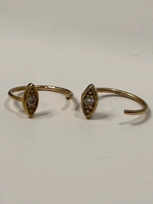 Guldiga hoops med kristalldetalj från Handcrsfted Cph - Snygga hoops i guldton med öppen ringdesign och en avlång, ögonformad detalj framtill. I mitten sitter en liten klar kristall som ger extra glans. Handgjorda från Handcrsfted Cph i materialet argentum och nysilver. Perfekta för dig som gillar stilrena och trendiga accessoarer.