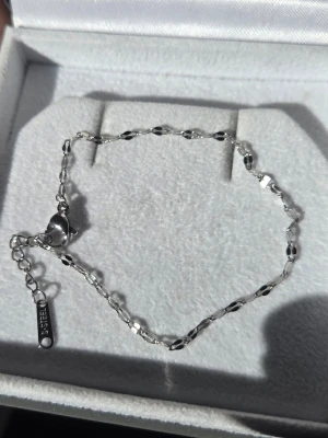 Silverfärgat armband i rostfritt stål - Snyggt och stilrent handgjort armband i silverfärgat rostfritt stål med små, blanka länkar i en tvistad design. Justerbar längd med karbinlås och bricka märkt 'S.STEEL'. Perfekt för dig som gillar minimalistiska smycken med en modern touch.