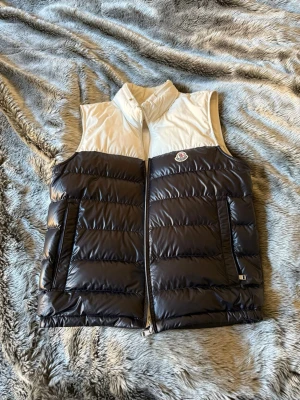 Moncler väst - Snygg dunväst från Moncler i mörkblått och vitt med hög krage och dragkedja framtill. Västen har Moncler-logga på bröstet och två fickor med dragkedja. Tillverkad i glansigt material och fylld med dun för extra värme. Perfekt för lager-på-lager-stil.