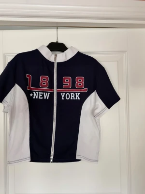Blåvit croppad zip-top med tryck - Sportig croppad T-shirt i mörkblått och vitt med två dragkedjor framtill. Tryck med '1898 New York' i rött och vitt på bröstet. Kortärmad och med hög krage och lite strävt material. Aldrig använd.