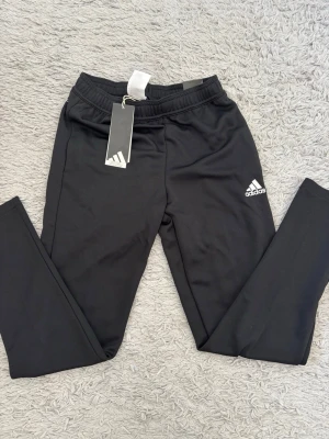 Svarta leggings från Adidas - Snygga svarta leggings från Adidas med klassisk logga på låret. De har en elastisk midja och är tillverkade i ett stretchigt material som passar perfekt till träning eller chill. Diskret Adidas-text på baksidan vid höften. Superskön passform och stilren look.