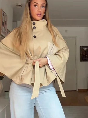 Beige trenchcoat med knytband - Stilren beige trenchcoat med vida ärmar och knytband i midjan. Jackan har hög krage och stora svarta knappar framtill. Materialet ser ut att vara bomull eller en bomullsblandning. Perfekt för dig som vill ha en trendig och avslappnad look. Storlek XS-S
